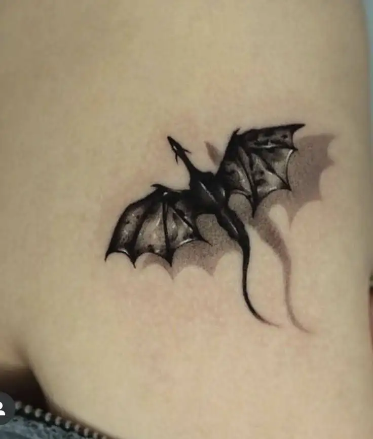 A small, detailed black dragon tattoo with its wings spread, casting a shadow on the skin.