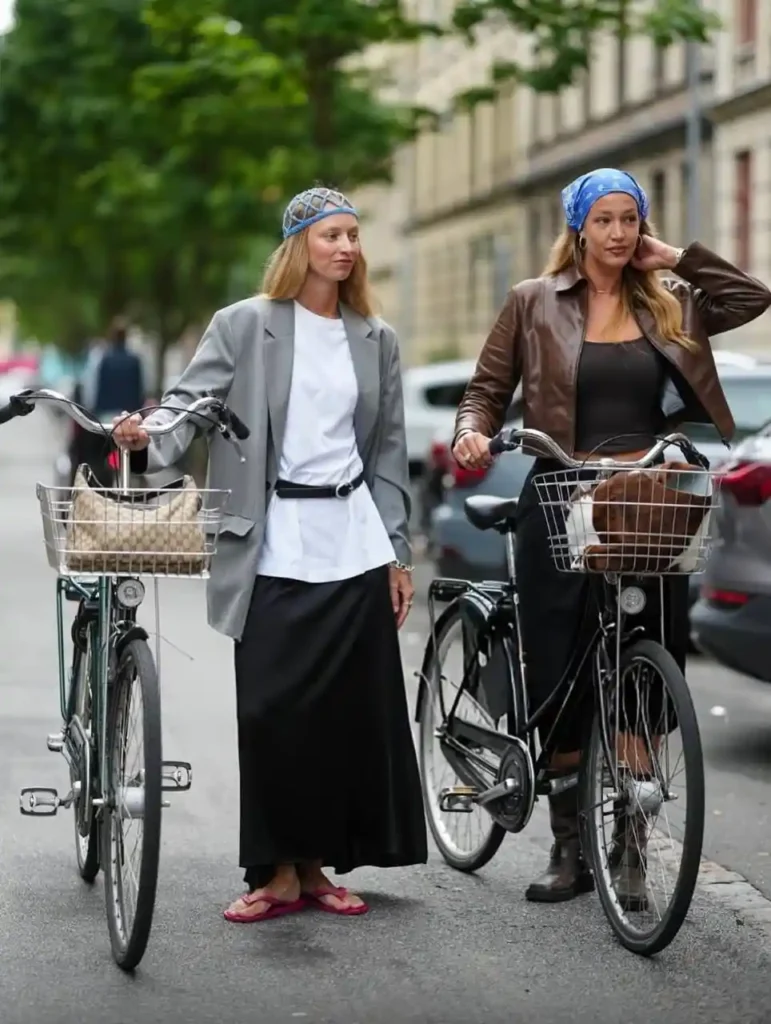 Two women in stylish streetwear with headscarves, walking beside bicycles on a city street