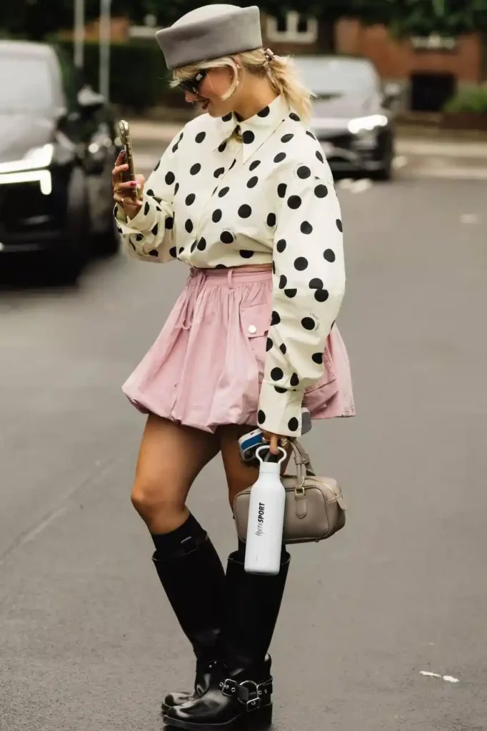 Street style look featuring a woman in a polka dot shirt, pink skirt, knee-high boots, and a beret, holding a phone and water bottle