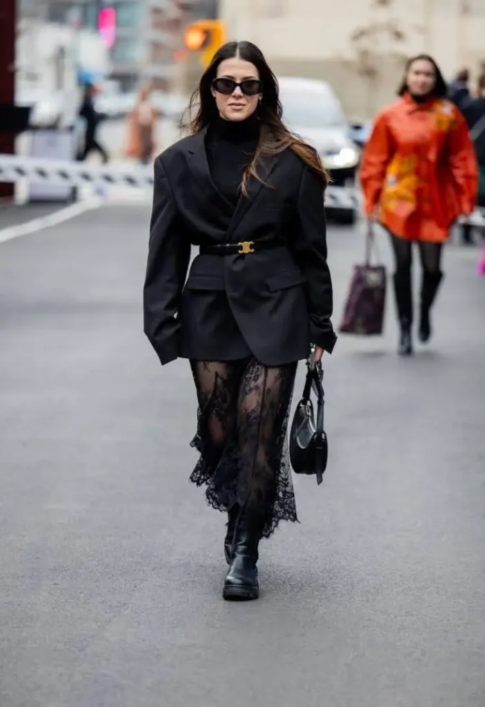 A woman wearing an all-black outfit with a blazer, a black turtleneck, and a sheer lace skirt.