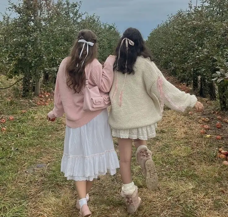 Two women in coquette aesthetic walking through an apple orchard.