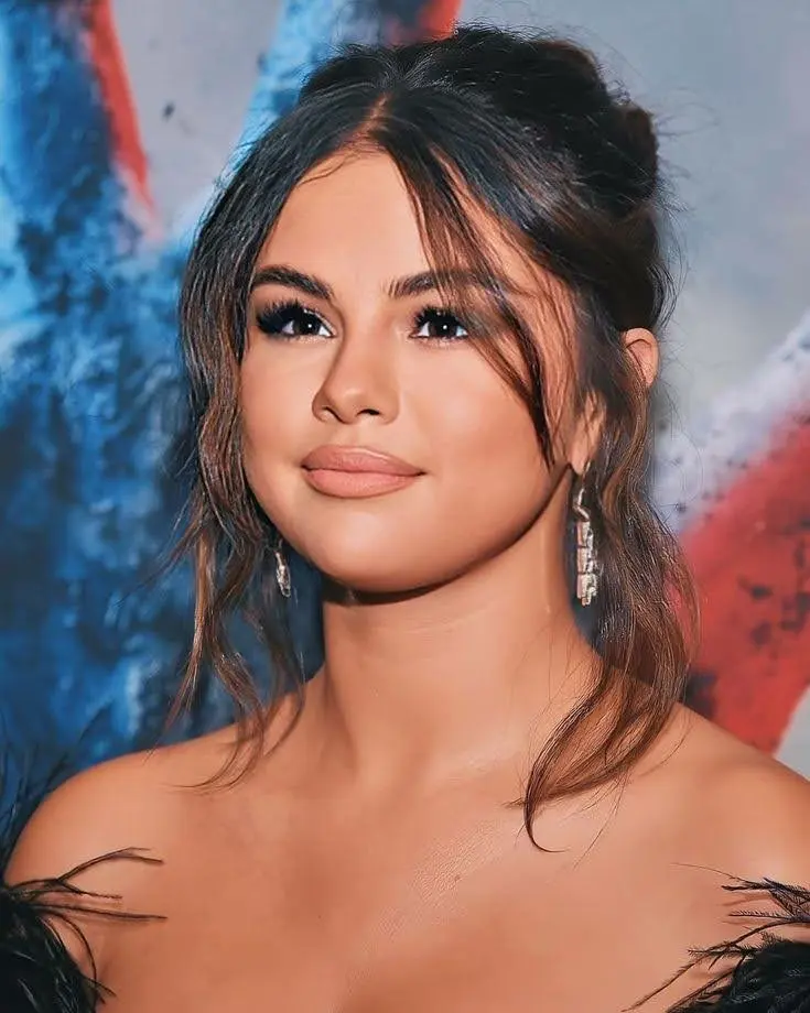 A close-up of Selena Gomez with her hair in a loose bun, a black off-the-shoulder dress, and drop earrings.