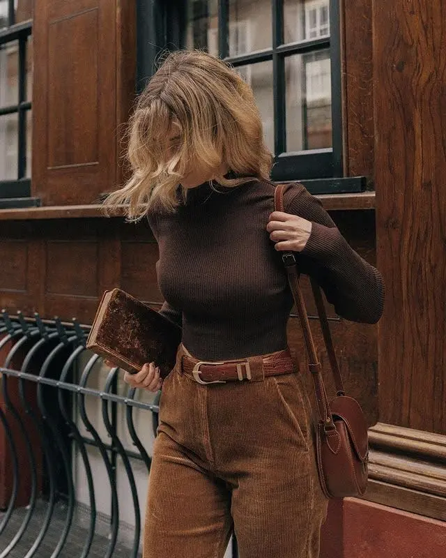 Woman in a dark academia outfit: a brown turtleneck, corduroy pants, and a leather satchel.