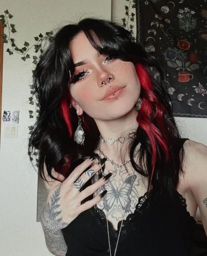 A young woman with dark hair, red highlights, tattoos, and piercings, embodying the e-girl aesthetic.