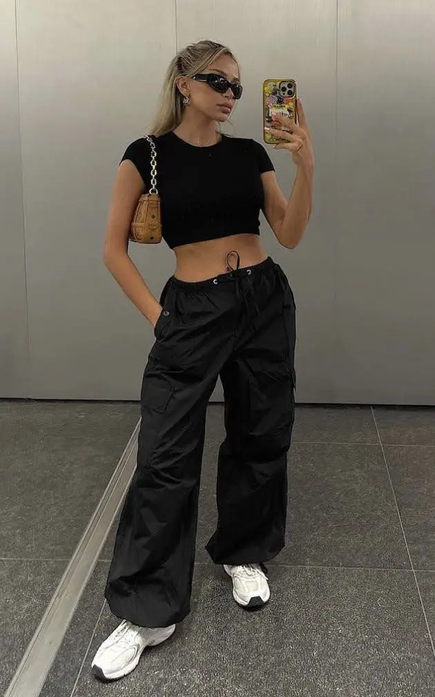 A young woman wearing a black cropped t-shirt and wide-leg black pants, taking a selfie.