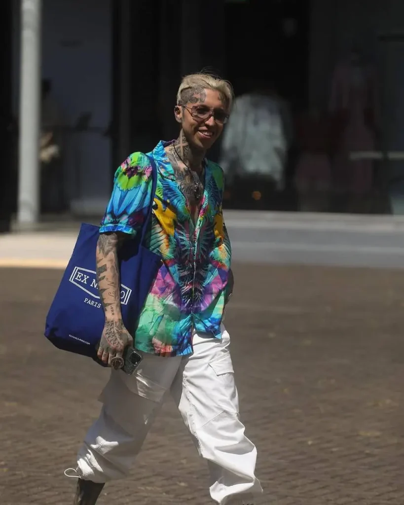 A stylish person with tattoos and dyed hair smiles while wearing a colorful button-down shirt.