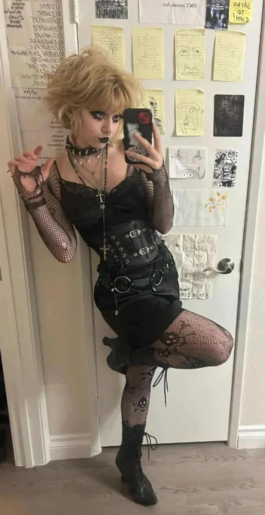 A stylish girl with stylish trad goth dress standing in front of mirror taking a mirror selfie.